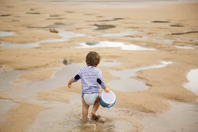 enfant plage Un enfant (environ 5-7 ans) est assis sur le sable fin d'Utah Beach, concentré à construire un grand château de sable avec un seau et une pelle, un soleil doux de juin illumine la scène.